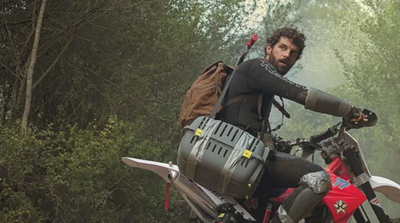 A man with a beard and curly hair rides a red dirt bike through a forest, wearing a black long-sleeve shirt, a brown…