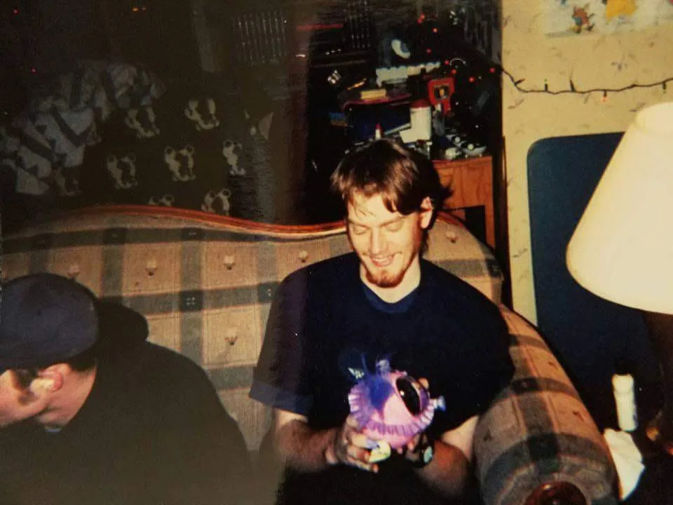 A grainy, low-light photo shows a man with a beard and dark t-shirt sitting on a plaid couch, smiling while holding a purple…