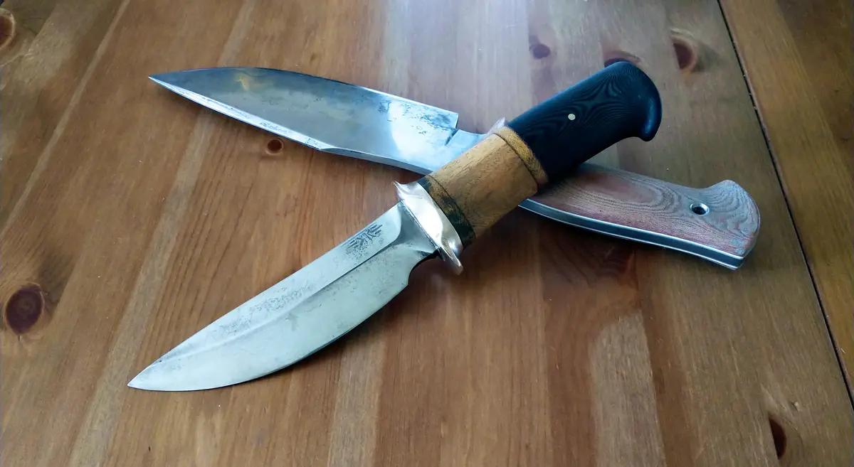 Two knives lie crossed on a wooden surface; one has a black textured handle and wooden bolster, the other has a…