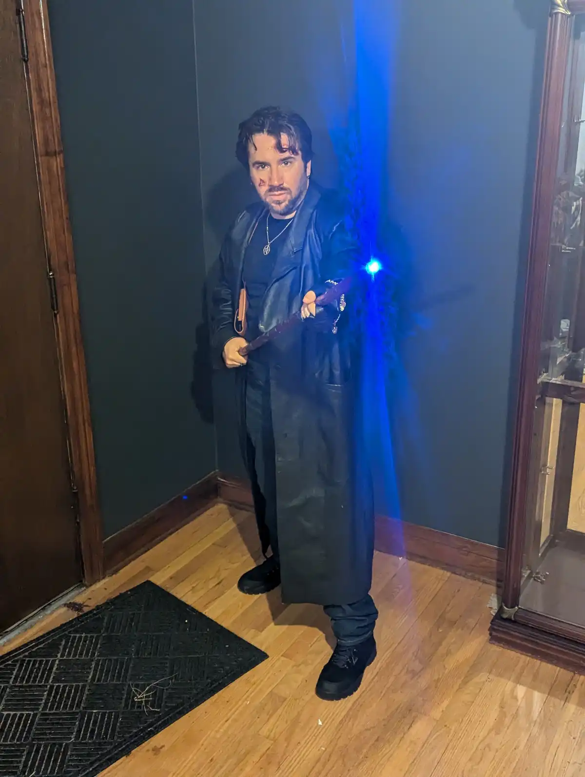 A man in a dark, long coat and black boots stands indoors on hardwood floors, holding a flashlight that emits a bright blue…