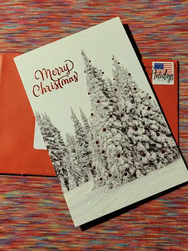 A white Christmas card with 'Merry Christmas' in copper script, featuring glittery snow-covered pine trees with red circular…