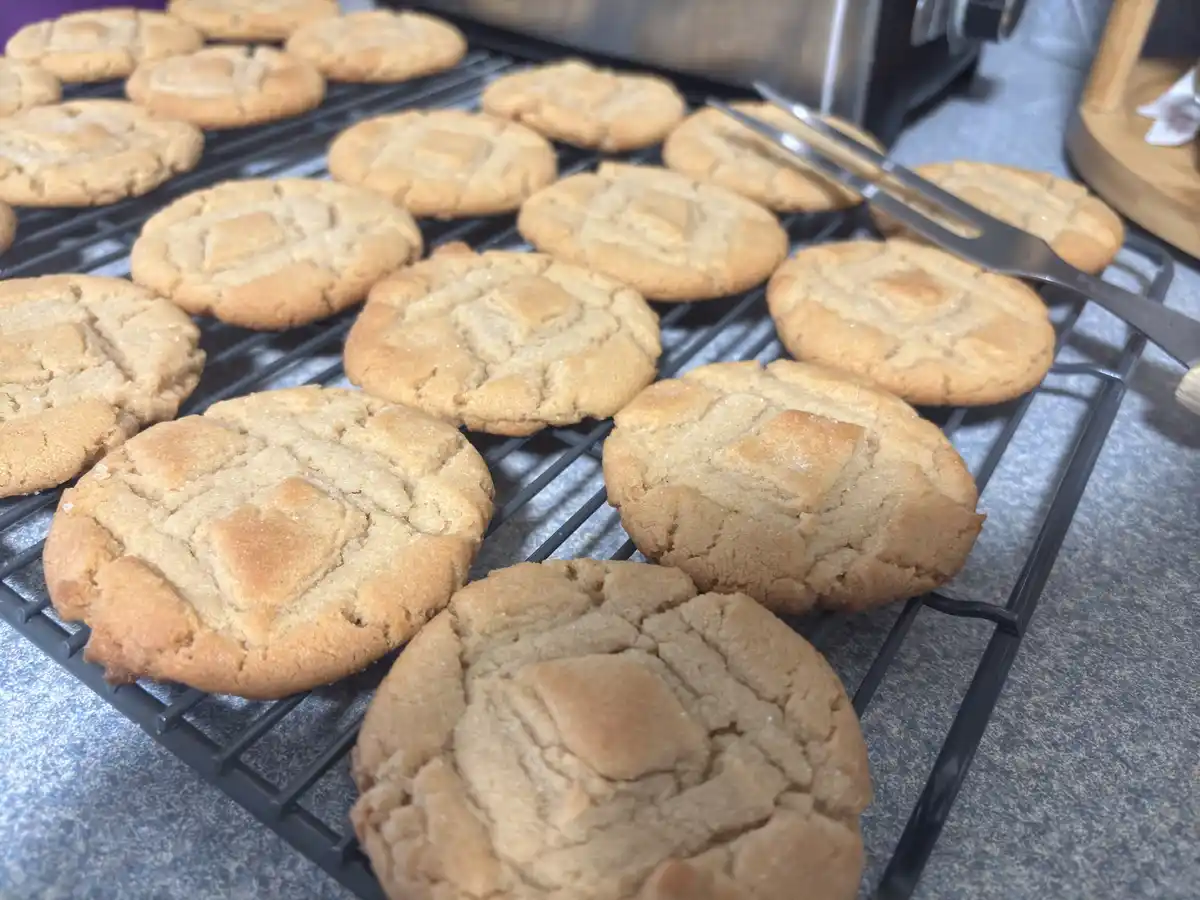 A close-up photo shows numerous freshly baked peanut butter cookies with a classic criss-cross pattern on a black wire…