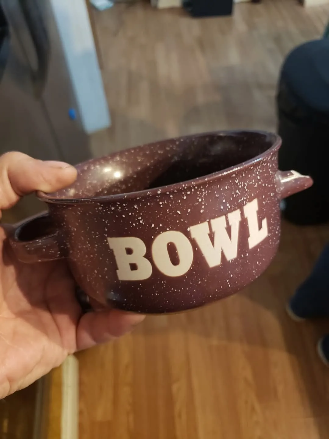 A hand holds a brown speckled ceramic bowl with the word 'BOWL' printed in large white letters on its side. The bowl has two…