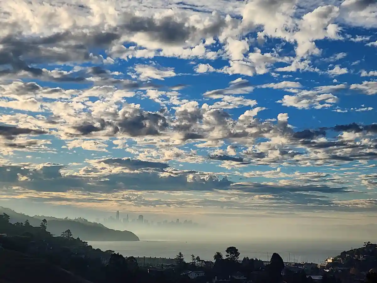 A scenic photo shows a vast blue sky filled with scattered white and gray clouds above a hazy coastal view. In the distance…