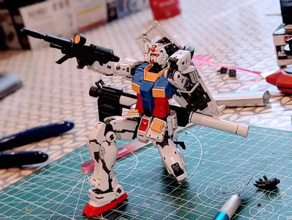 A white, blue, and red Gundam model kit stands on a green cutting mat surrounded by modeling tools and parts, holding a…