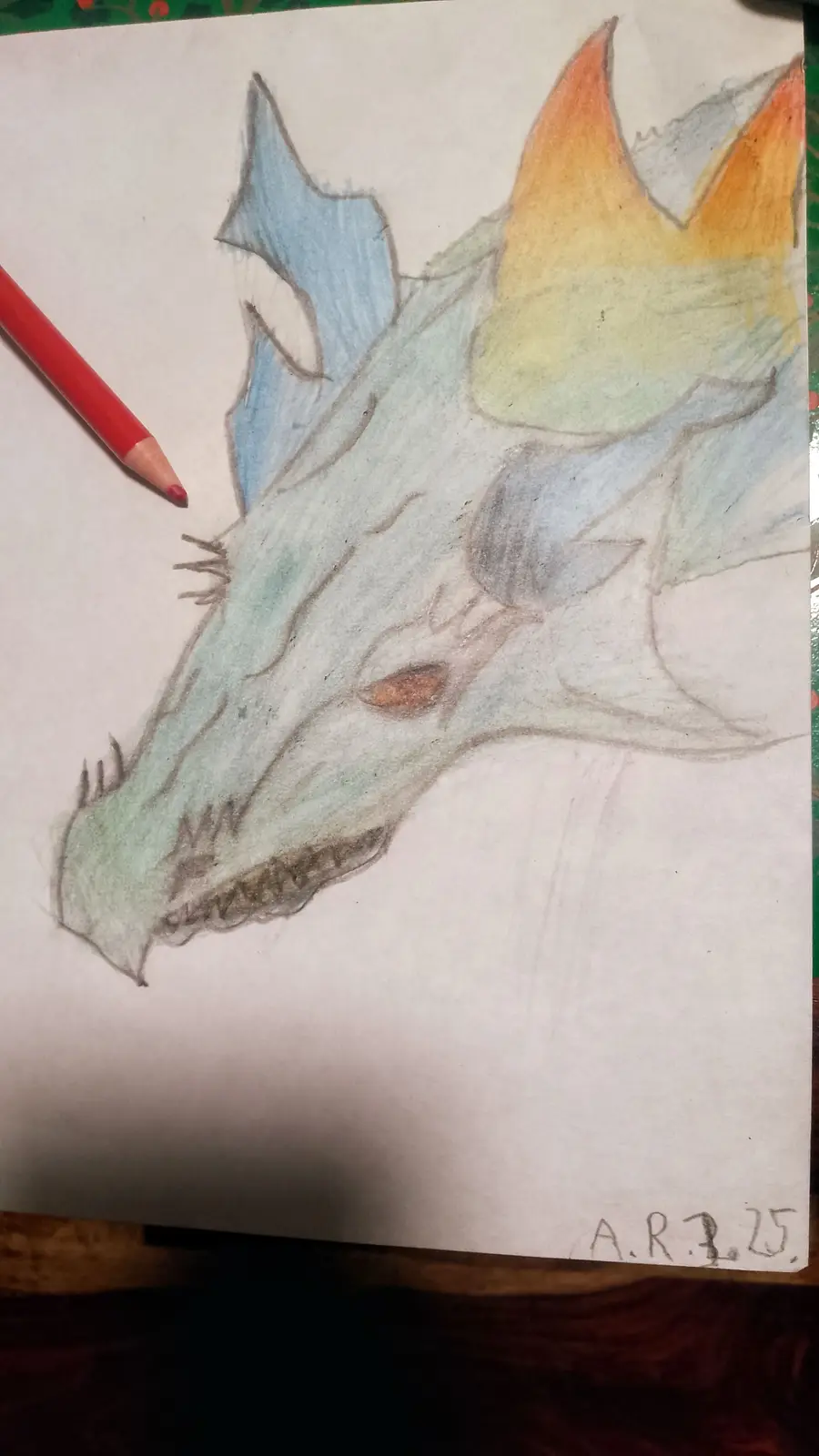 A hand-drawn colored pencil sketch of a dragon's head on white paper, featuring blue and green scales, orange eyes, and…