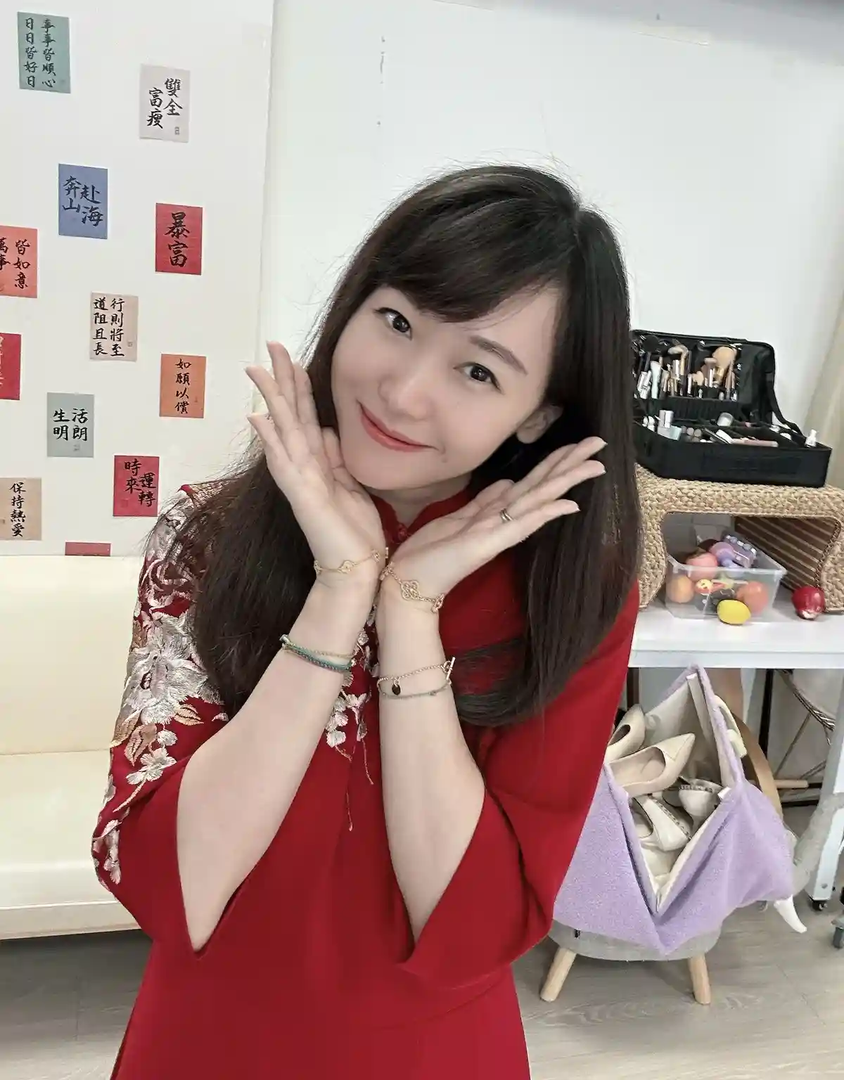 A woman in a red traditional-style dress smiles at the camera with her hands framing her face, standing in front of a wall…