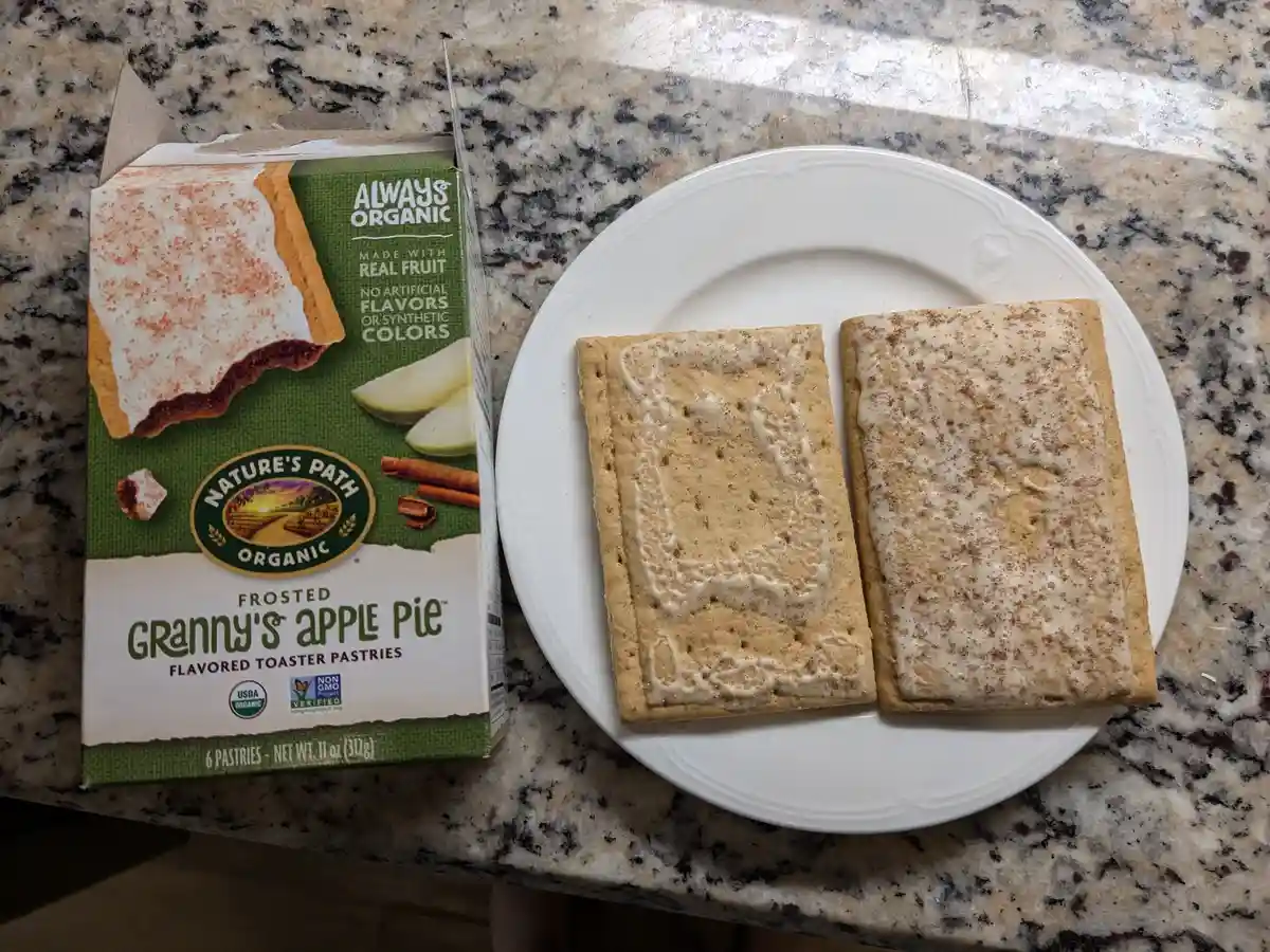 A box of Nature's Path Frosted Granny's Apple Pie toaster pastries sits next to a white plate holding two of the pastries…
