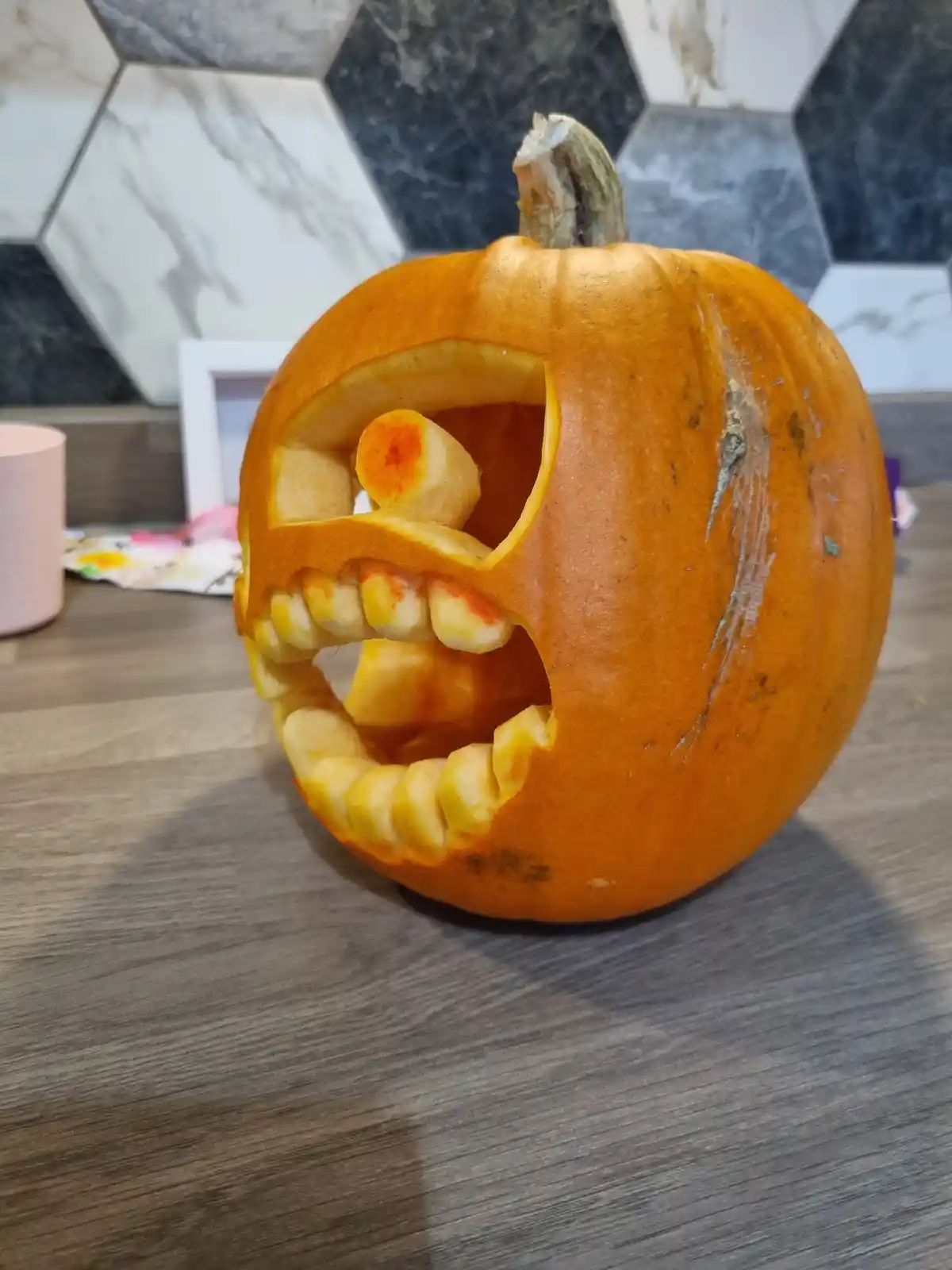 A close-up of an orange pumpkin carved with a wide, toothy grin, sitting on a wooden surface. The carved teeth are made from…