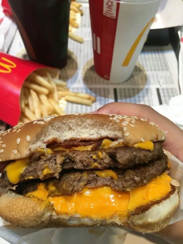 A hand holds a large, double-patty cheeseburger with melted cheese and sesame seed bun, partially eaten. In the background…