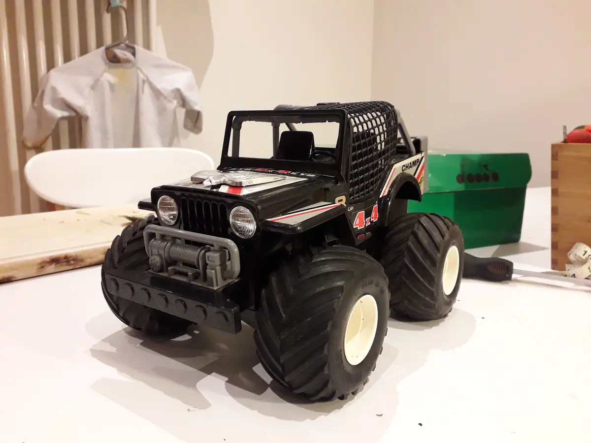 A black toy monster truck with oversized tires and a protective cage sits on a white table, with a white shirt hanging on a…
