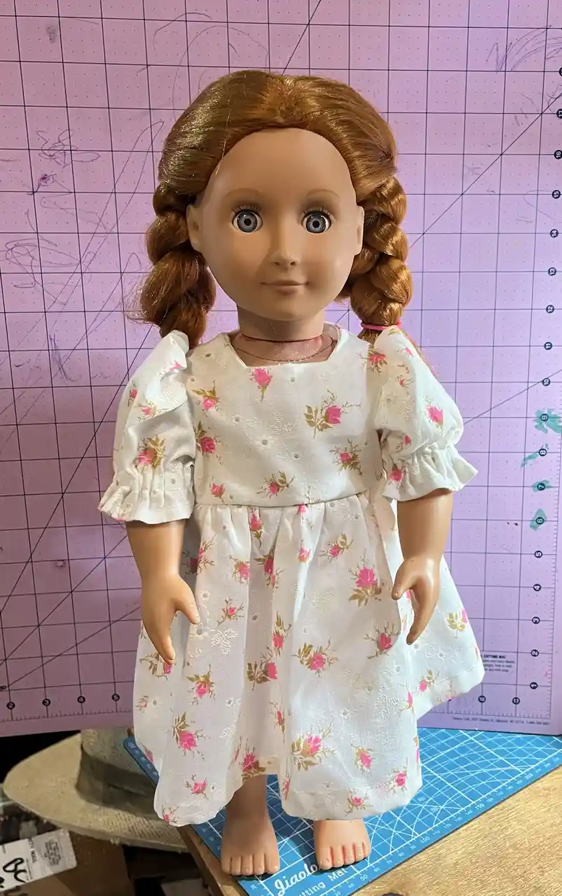 A doll with brown braided hair and large blue eyes wears a handmade white floral dress with pink and gold roses, standing on…