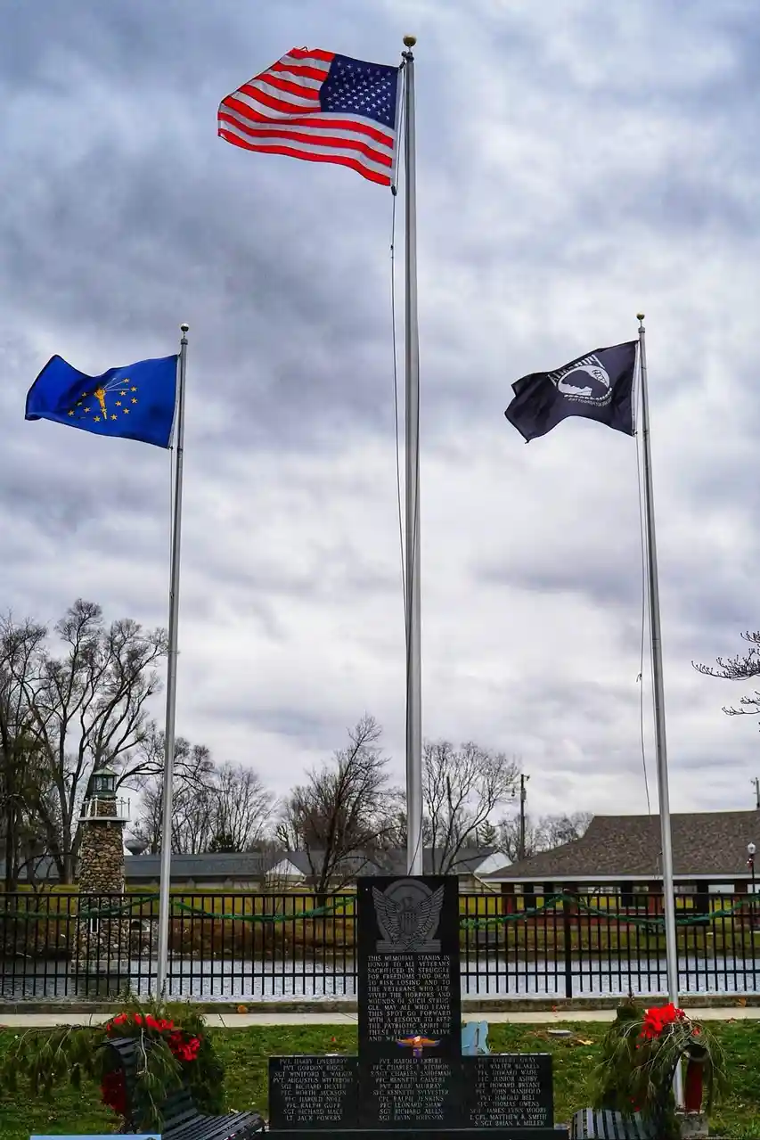 A memorial plaque with engraved names is centered between three flagpoles flying the American flag, the Alaska state flag…