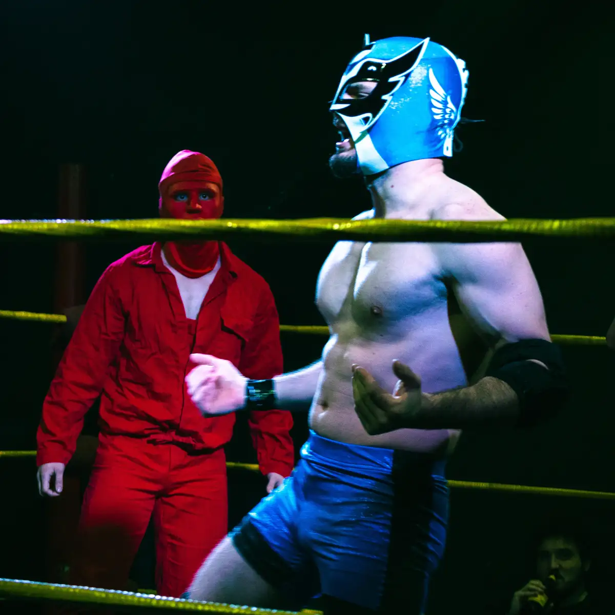 A wrestler in a blue mask and trunks stands in a ring, gesturing dismissively at another man in a red jumpsuit and head…