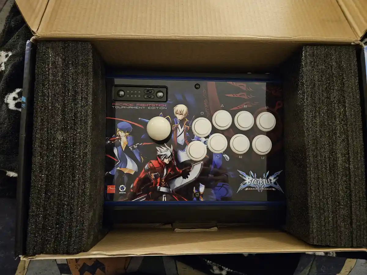 An arcade fightstick, branded 'BlazBlue: Continuum Shift' and labeled 'Arcade Fightstick Tournament Edition', is shown…