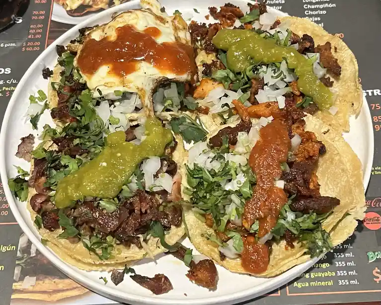 A plate of four tacos topped with shredded meat, chopped onions, cilantro, and either red or green salsa, one with a fried…
