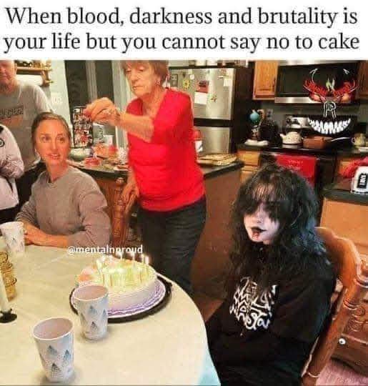 A person with black face paint and long black hair sits at a kitchen table with a birthday cake in front of them, looking…