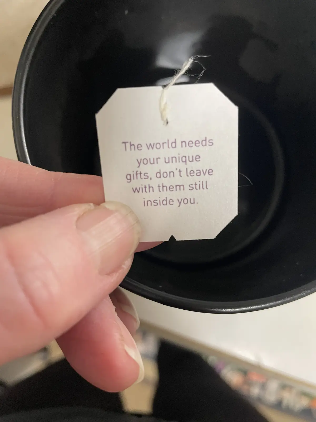 A close-up photo shows a hand holding a white, octagonal tea bag tag with purple text against the dark interior of a black…