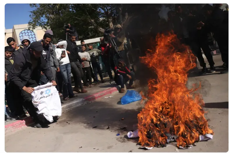 A man kneels beside a large fire burning a white shirt with a blue Star of David and Arabic text. A crowd of men and boys…