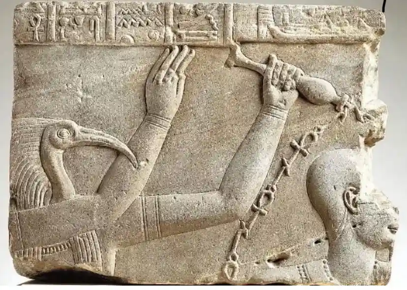 An ancient Egyptian stone relief carving depicts a bird-headed deity with human arms, one hand holding a staff and the other…