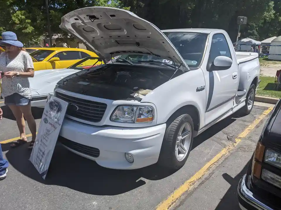 A white Ford F-150 Lightning pickup truck with its hood open is parked at an outdoor car show, displaying its engine. A…