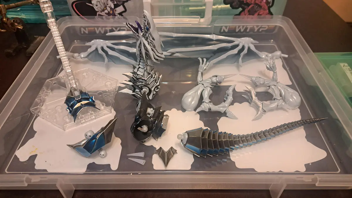 A partially assembled white and blue plastic dragon model sits in a clear plastic storage box alongside its disassembled…