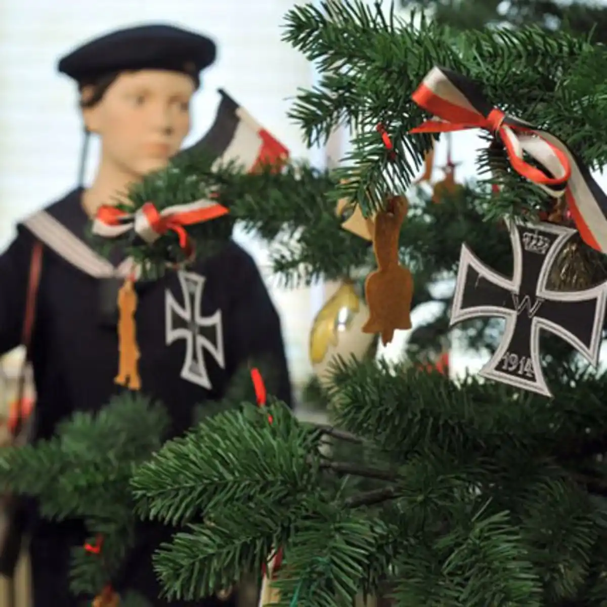 A close-up of a Christmas tree decorated with German Iron Cross medals and red-white-black ribbons, with a blurred mannequin…