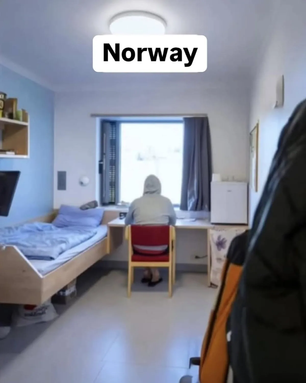 A person in a grey hoodie sits at a desk in a minimalist, brightly lit room with a single bed and a window, with the word…
