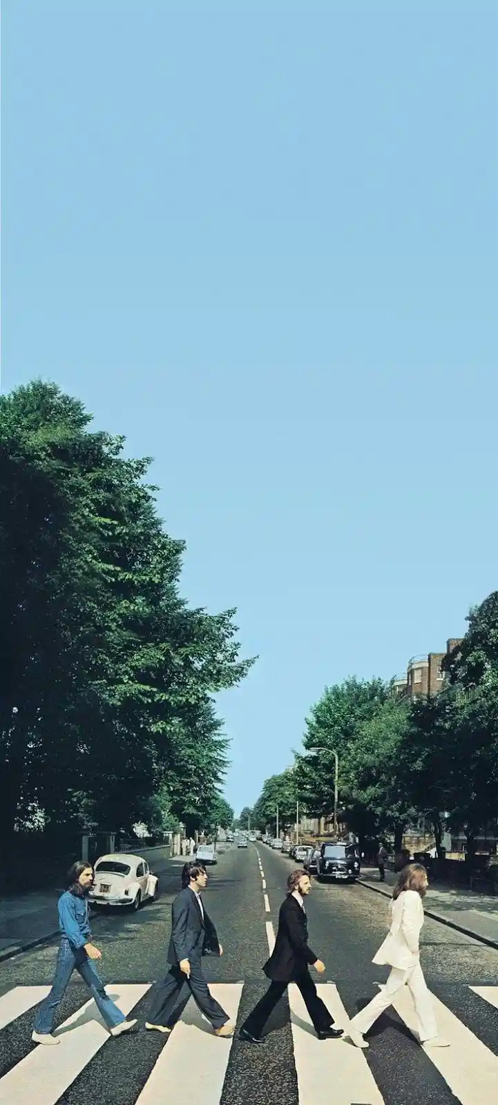 four men in suits and casual wear walk across a zebra crossing on a tree-lined street under a clear blue sky, with cars…