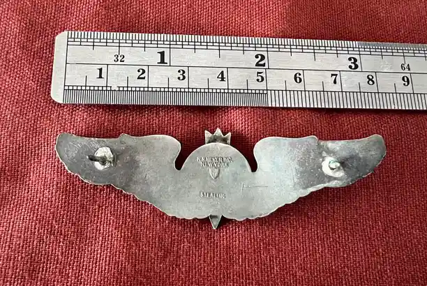 A silver, wing-shaped brooch with a crown on top and two pin-backs lies on a red textured surface next to a metal ruler…