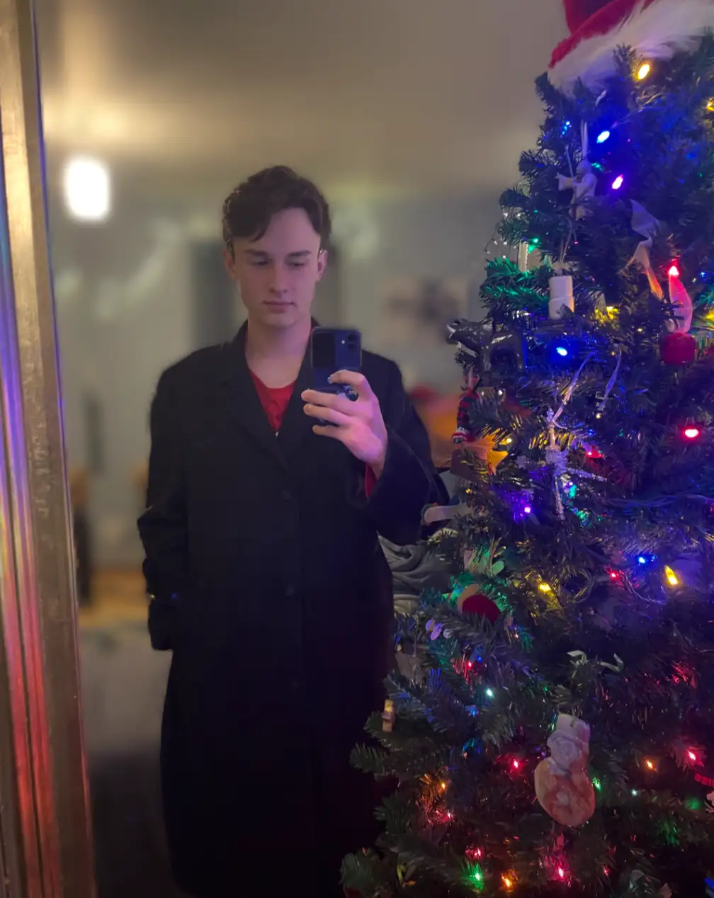 A young man in a black coat stands in front of a brightly lit, decorated Christmas tree, taking a mirror selfie with his…