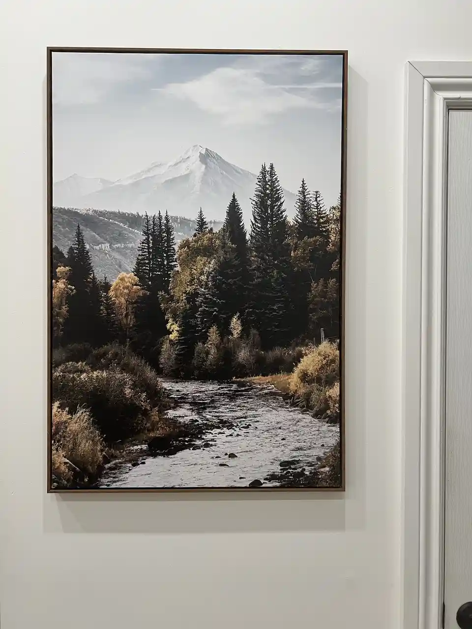 A framed photograph of a mountainous landscape with a river, trees, and snow-capped peaks hangs on a white wall next to a…