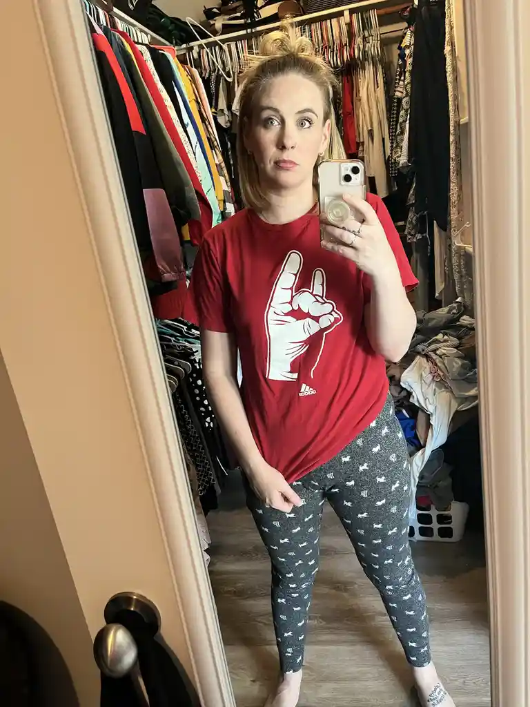A woman with blonde hair in a messy bun stands in a cluttered walk-in closet, taking a mirror selfie with her phone. She…
