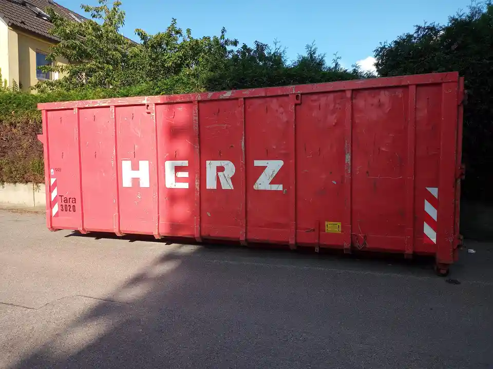 A large red dumpster with the word 'HERZ' (German for 'heart') painted in white letters on its side sits on an asphalt road…