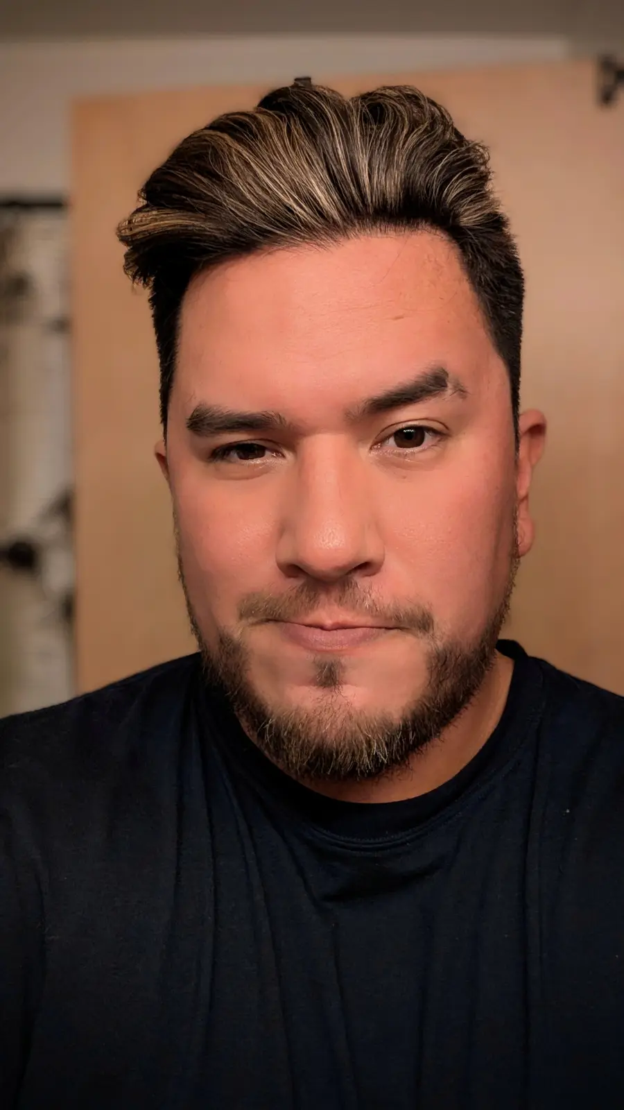 A man with styled hair and a goatee stares directly at the camera with a neutral expression. He wears a dark t-shirt and is…