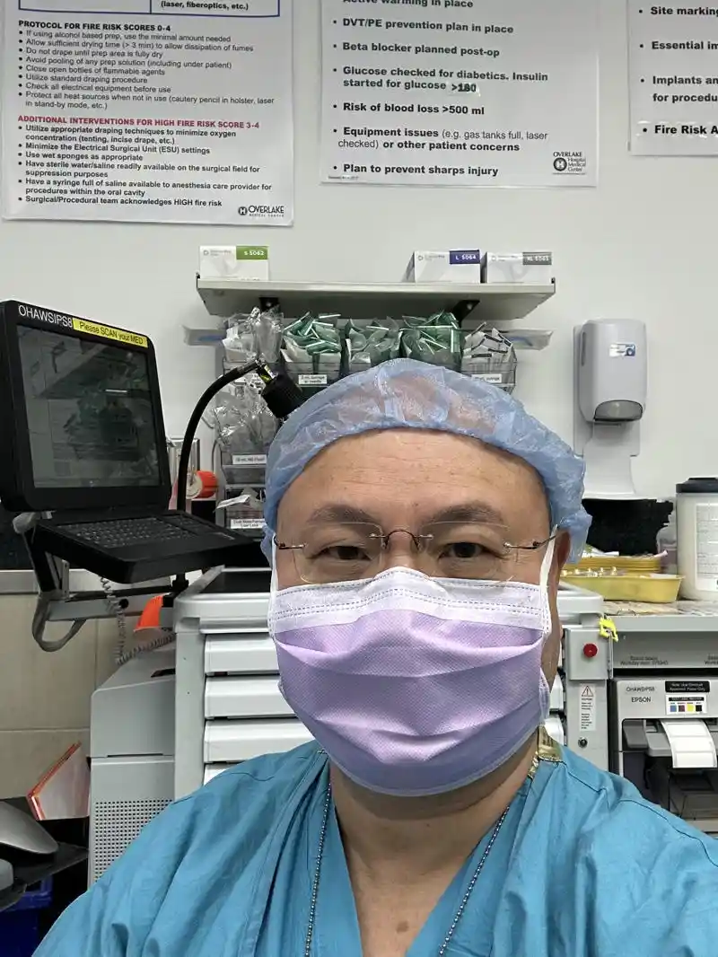 A person in blue surgical scrubs, a light blue surgical cap, and a light purple surgical mask looks directly at the camera…