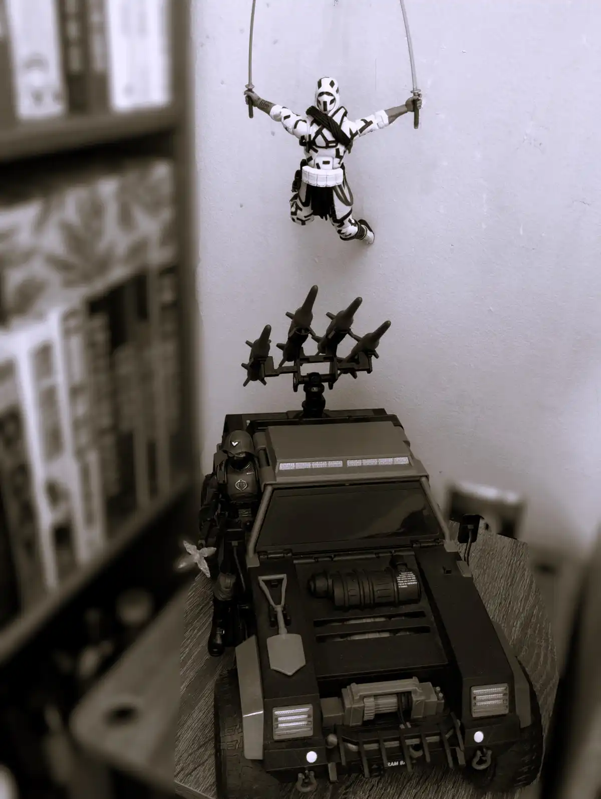 A sepia-toned photo shows a white and black ninja-style action figure suspended by strings from a wall above a black armored…