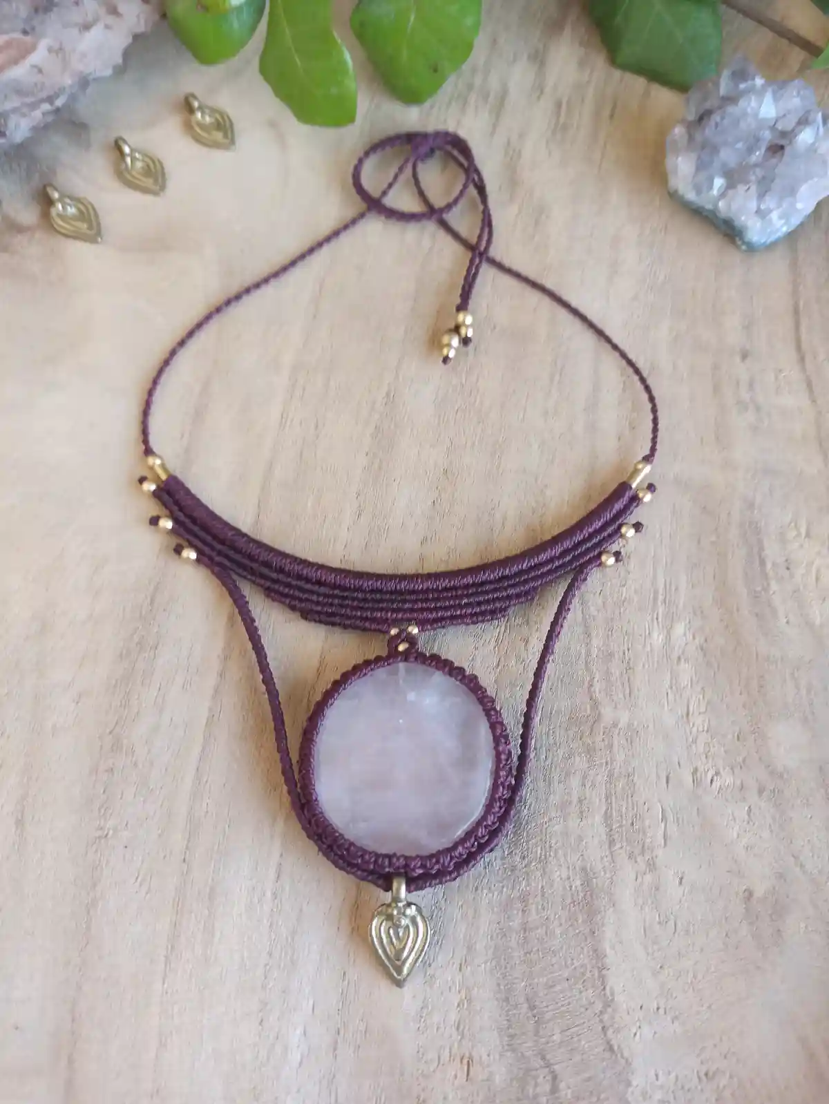 A handmade purple macrame necklace with a central rose quartz pendant and a small heart charm hangs on a wooden surface…