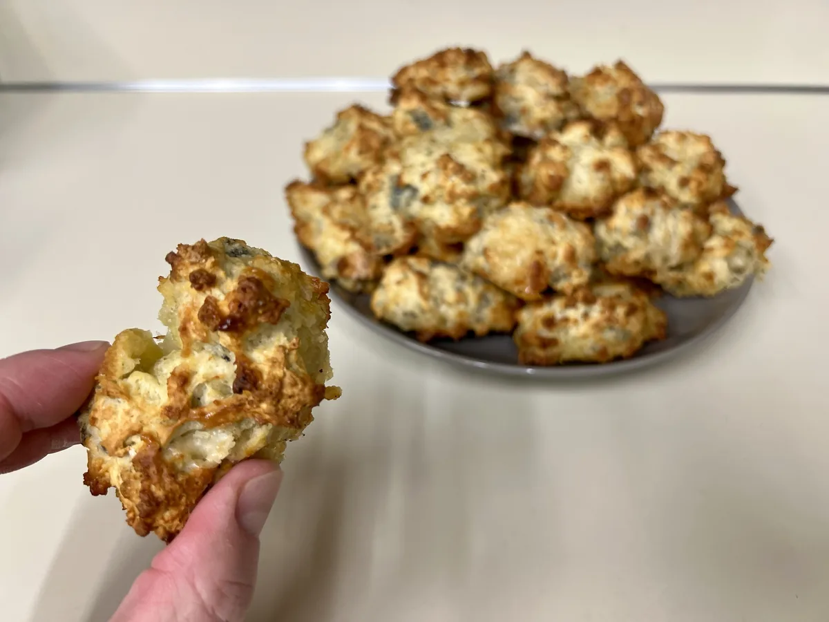 A hand holds up a broken, golden-brown baked good, revealing a gooey, cheesy interior, with a plate full of similar items…