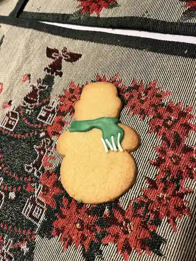A snowman-shaped sugar cookie with green icing scarf and white icing stick arms rests on a red, black, and beige embroidered…