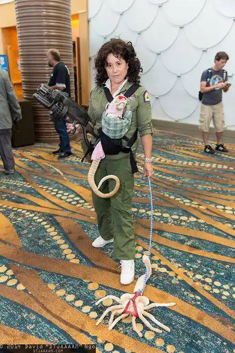 A woman dressed in a Ghostbusters uniform, holding a proton pack and a baby in a carrier, walks with a toy ghost on a leash…