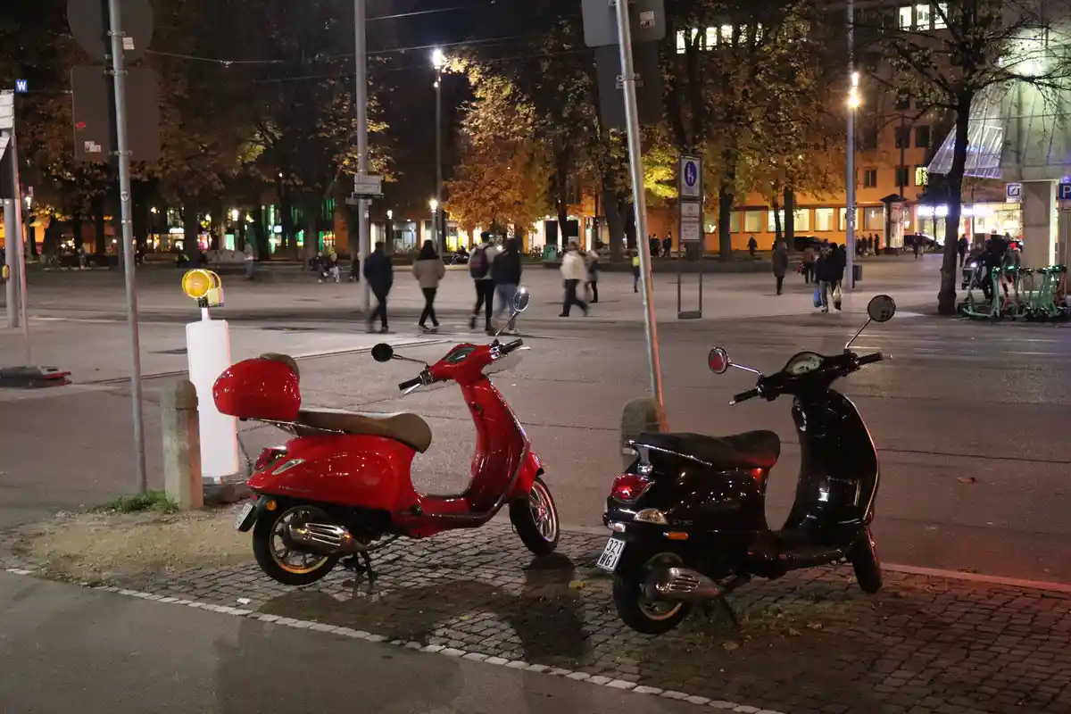 Two scooters, one red and one black, are parked side-by-side on a cobblestone sidewalk at night in an urban setting with…