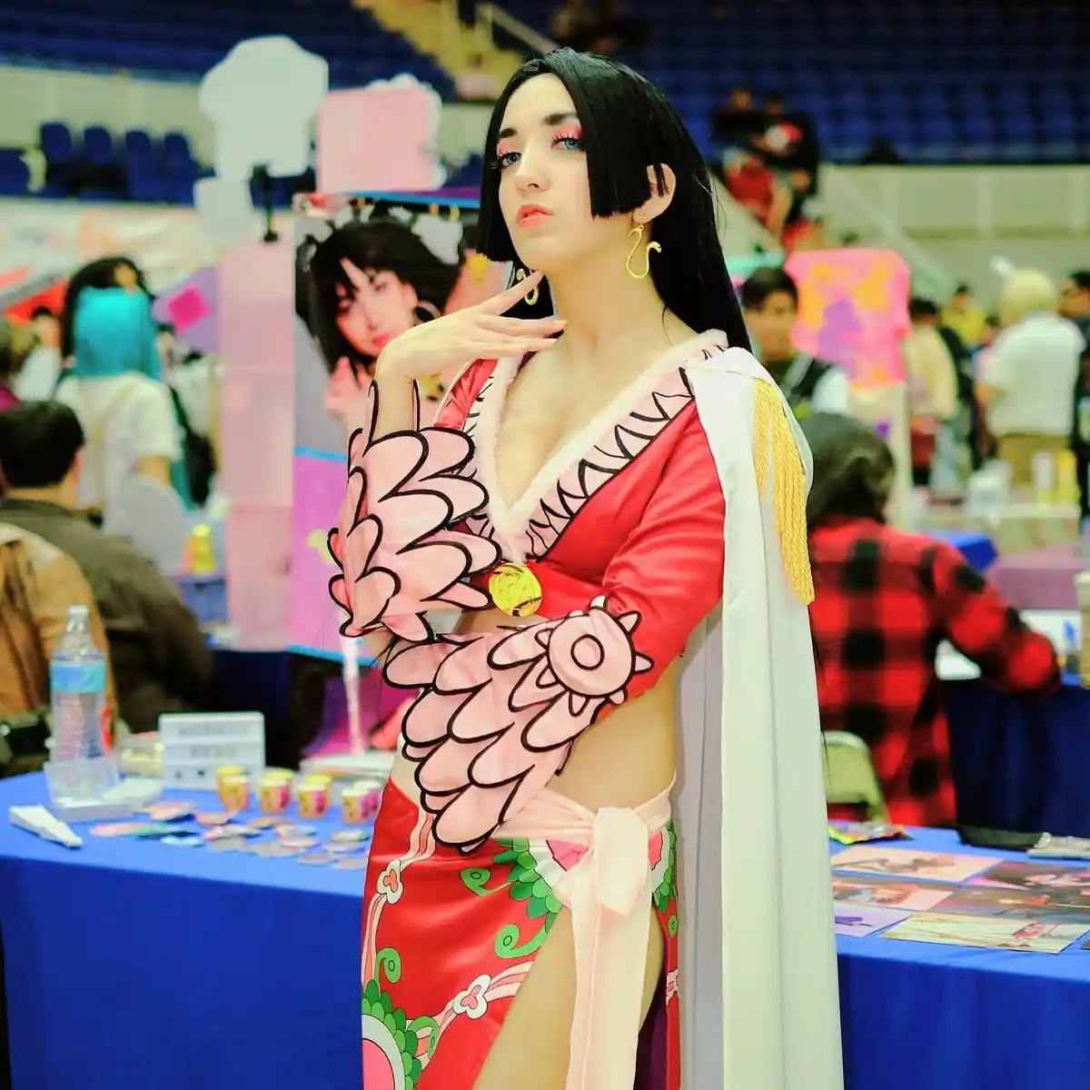 A cosplayer at a convention poses in a detailed red and pink costume with a white cape, holding her hand to her chin in a…