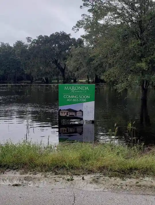 A real estate sign for 'Maronda Homes' advertising a new development is partially submerged in floodwater, with the text…