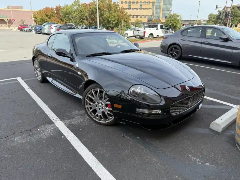A glossy black Maserati sports car is parked in a public asphalt lot, angled toward the viewer, with other vehicles and…