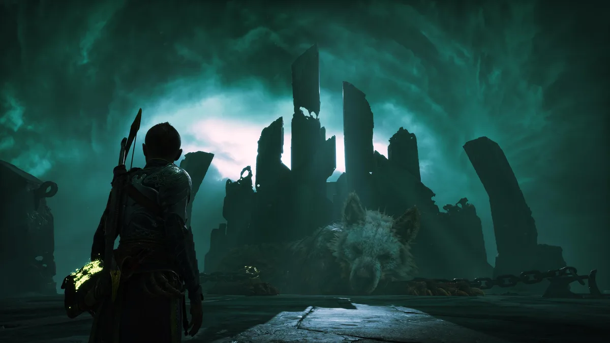 A character from the video game 'God of War' stands in a dark, teal-lit ruin, looking at a chained, deceased wolf-like…