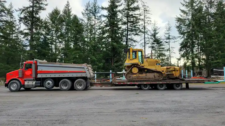 A red semi-truck with a dump bed is hauling a flatbed trailer carrying a large yellow Caterpillar D5H XL bulldozer. The…