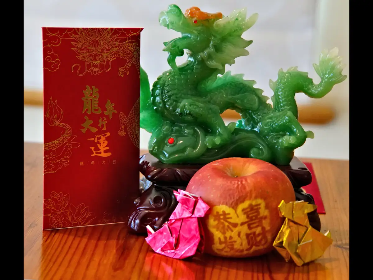 A still life of Chinese New Year decorations including a green jade dragon statue, a red envelope with gold dragon and text…