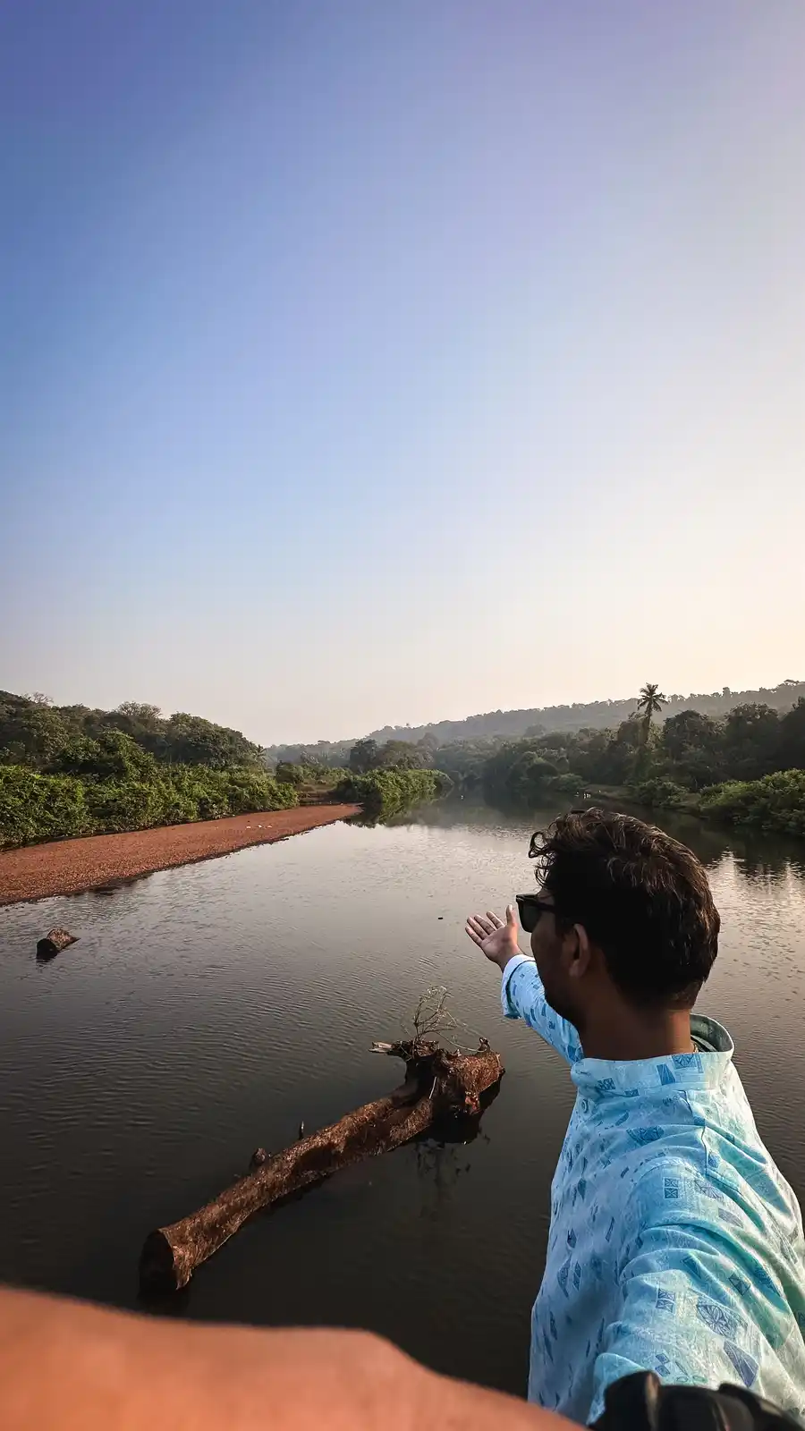 A man in a blue patterned shirt and sunglasses stands by a calm river, gesturing with his hand toward the water as if…