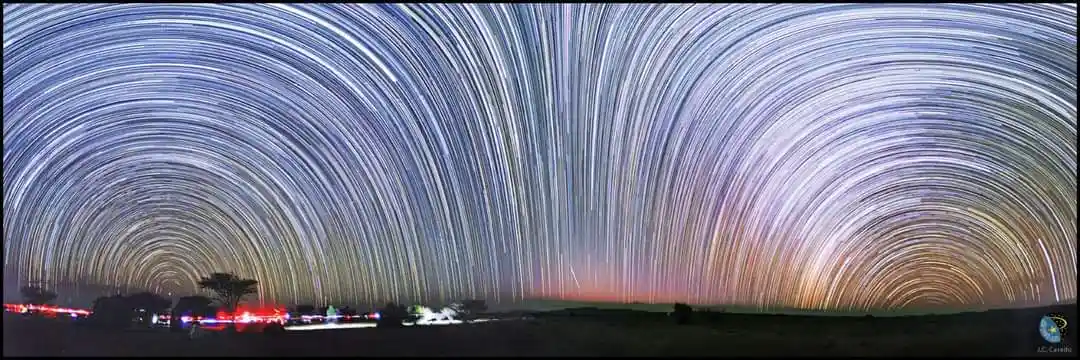 A long-exposure photograph shows sweeping arcs of star trails across a night sky, curving over a dark landscape with faint…